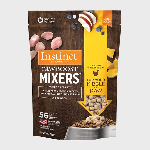 Dog food bag displays freeze-dried raw chicken mixers; scattered ingredients, text promotes grain-free, cage-free recipe; placed in kitchen-like setting.