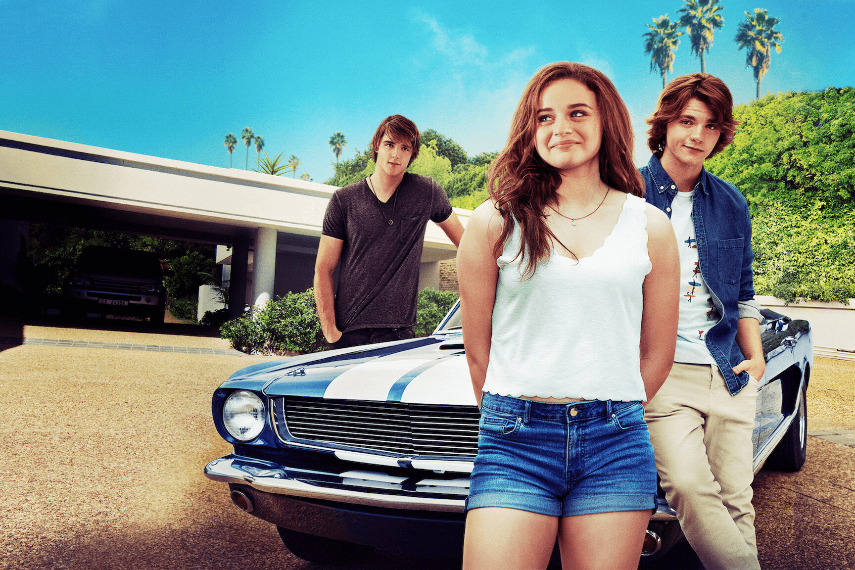Three people stand casually beside a classic car under a clear blue sky, with palm trees and a modern house in the background.