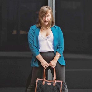 Person stands confidently, clutching a large black handbag, wearing a blue jacket over a white top, in an urban setting.
