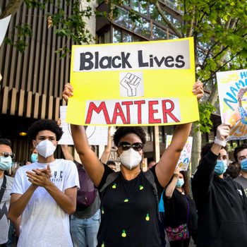 Protesters hold signs reading "Black Lives Matter" and "Say Her Name" in a city street, surrounded by trees and buildings, wearing masks.