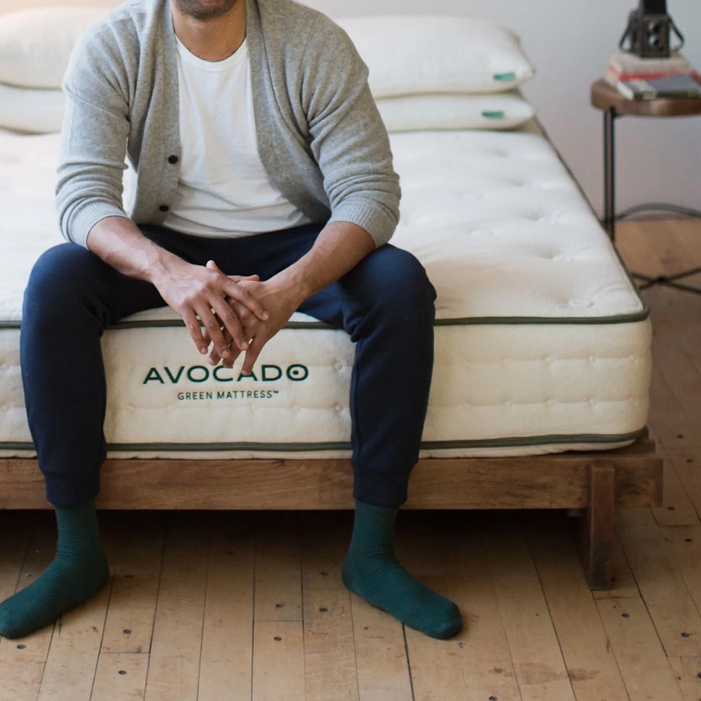 Person sitting on bed, hands clasped, wearing casual clothes and green socks; "Avocado Green Mattress" logo visible on bed. Wooden floor and side table nearby.
