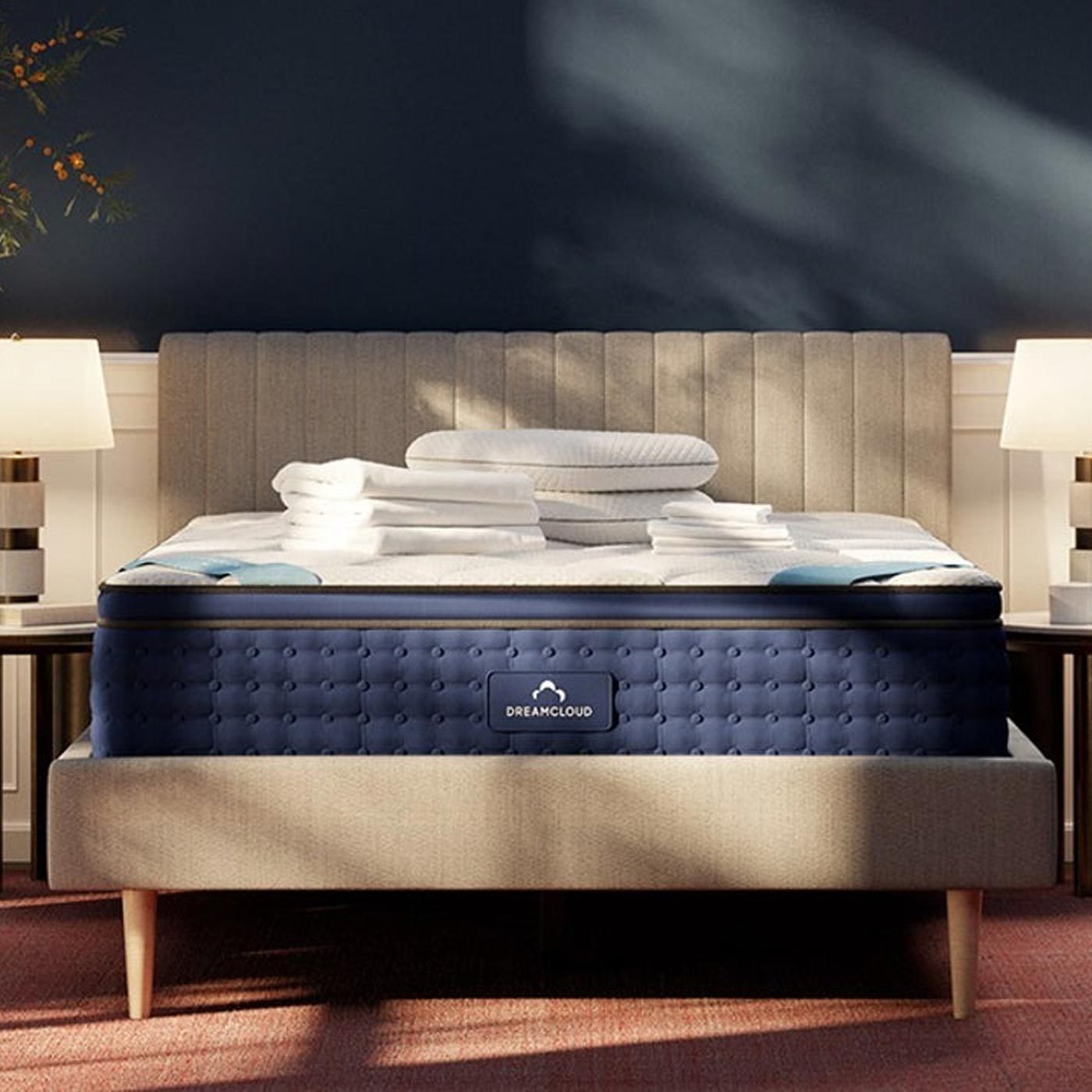 Bed with a neatly stacked pile of white linens; situated between two lamps in a softly lit, cozy bedroom. "DREAMCLOUD" is on the mattress.