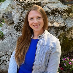 A woman in a blue shirt and denim jacket smiles, sitting in front of large moss-covered rocks with flowers nearby.