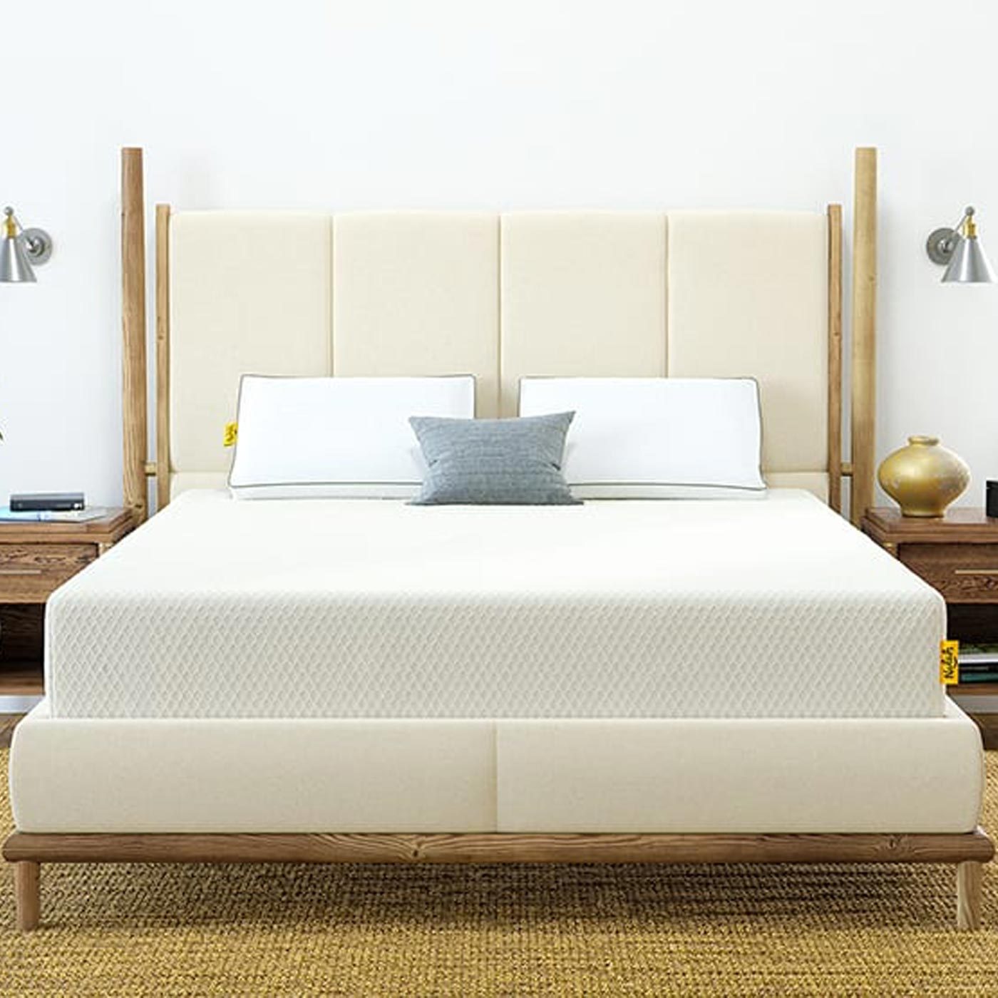 Bed with beige upholstery sits against a white wall, flanked by wooden side tables and wall-mounted lights, on a textured rug.