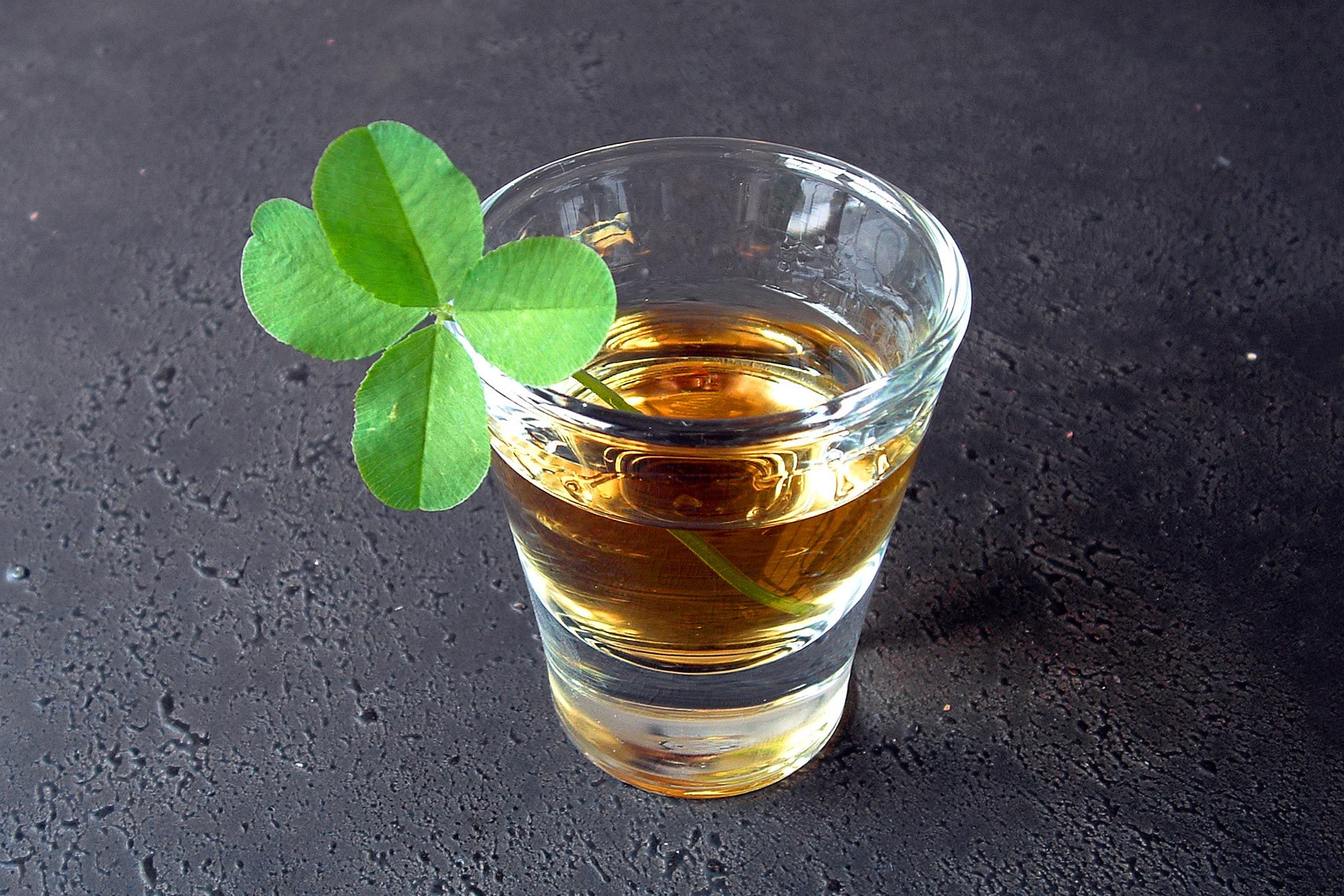 A glass of amber liquid sits on a dark surface, adorned with a green clover leaf.