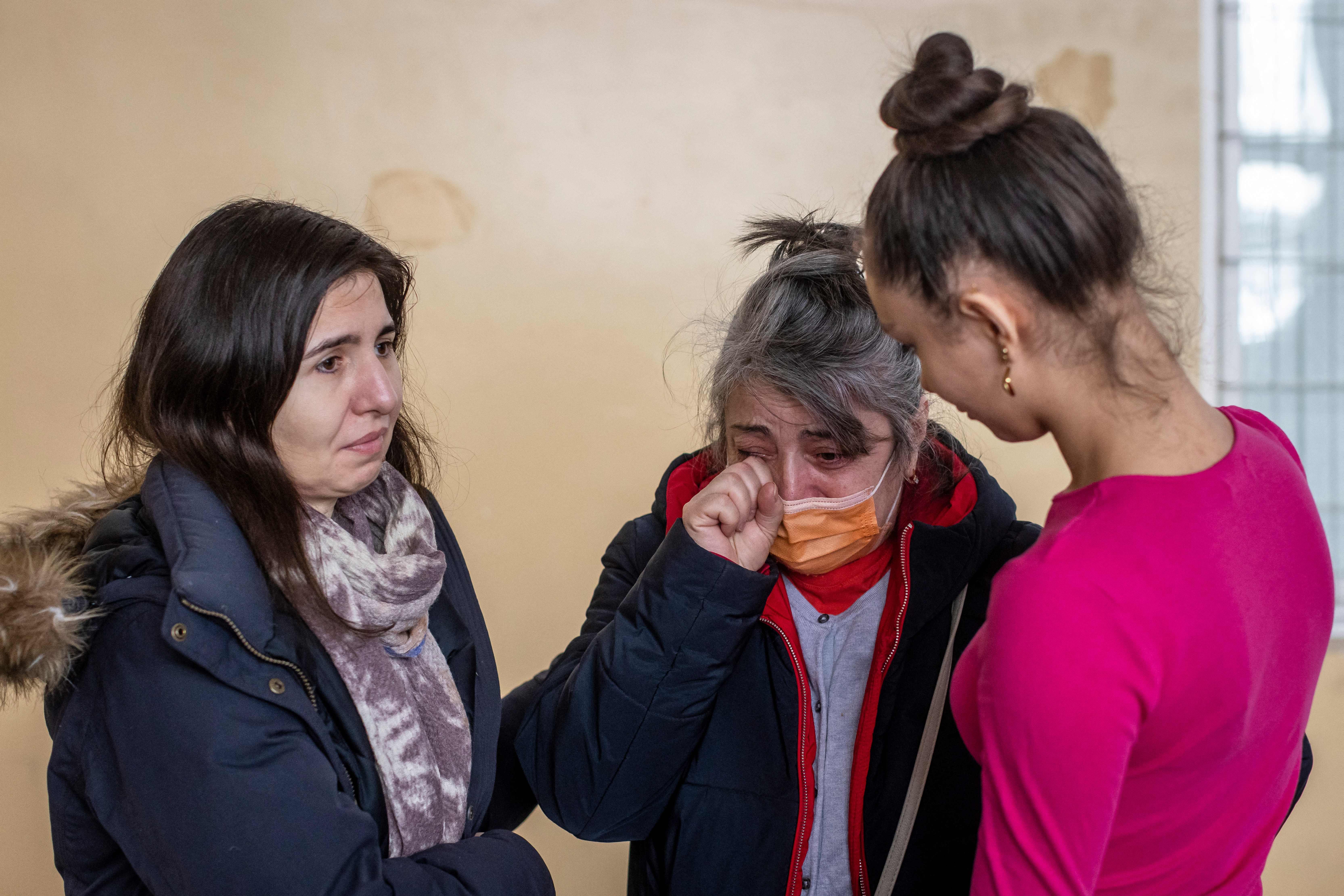 Three women stand closely; one wipes her eye while wearing a mask. They seem emotional in a warmly lit, indoor setting.