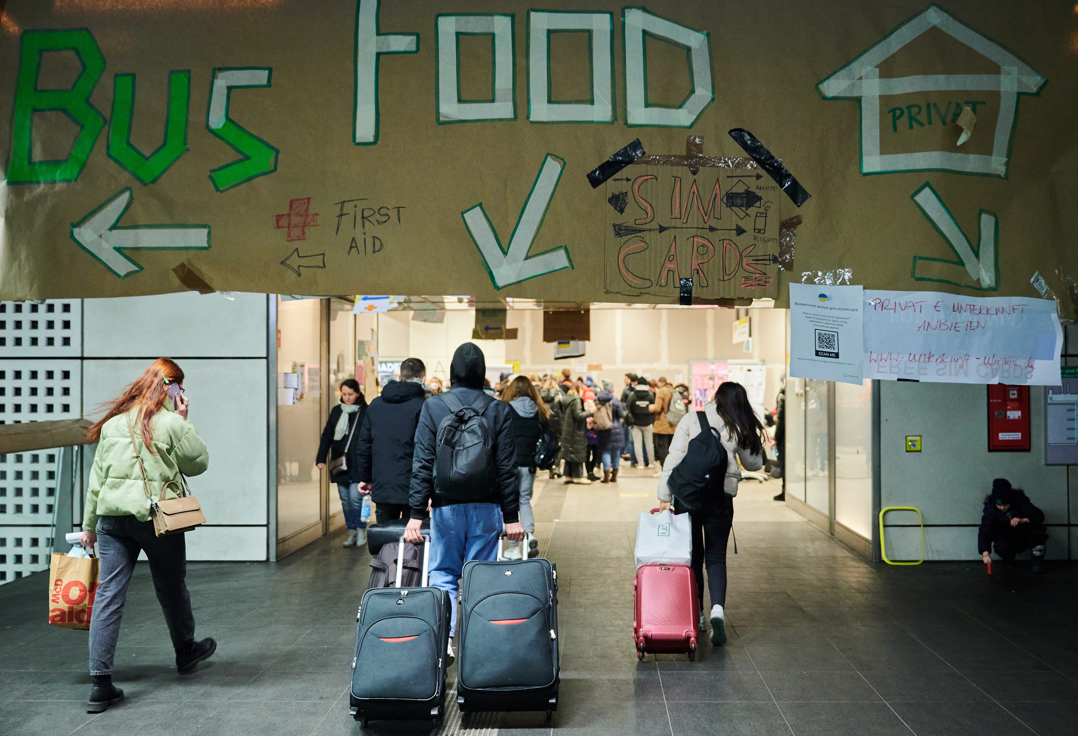People with luggage walk towards crowded indoor area; large sign overhead directs to buses, food, first aid, SIM cards, and private accommodations.