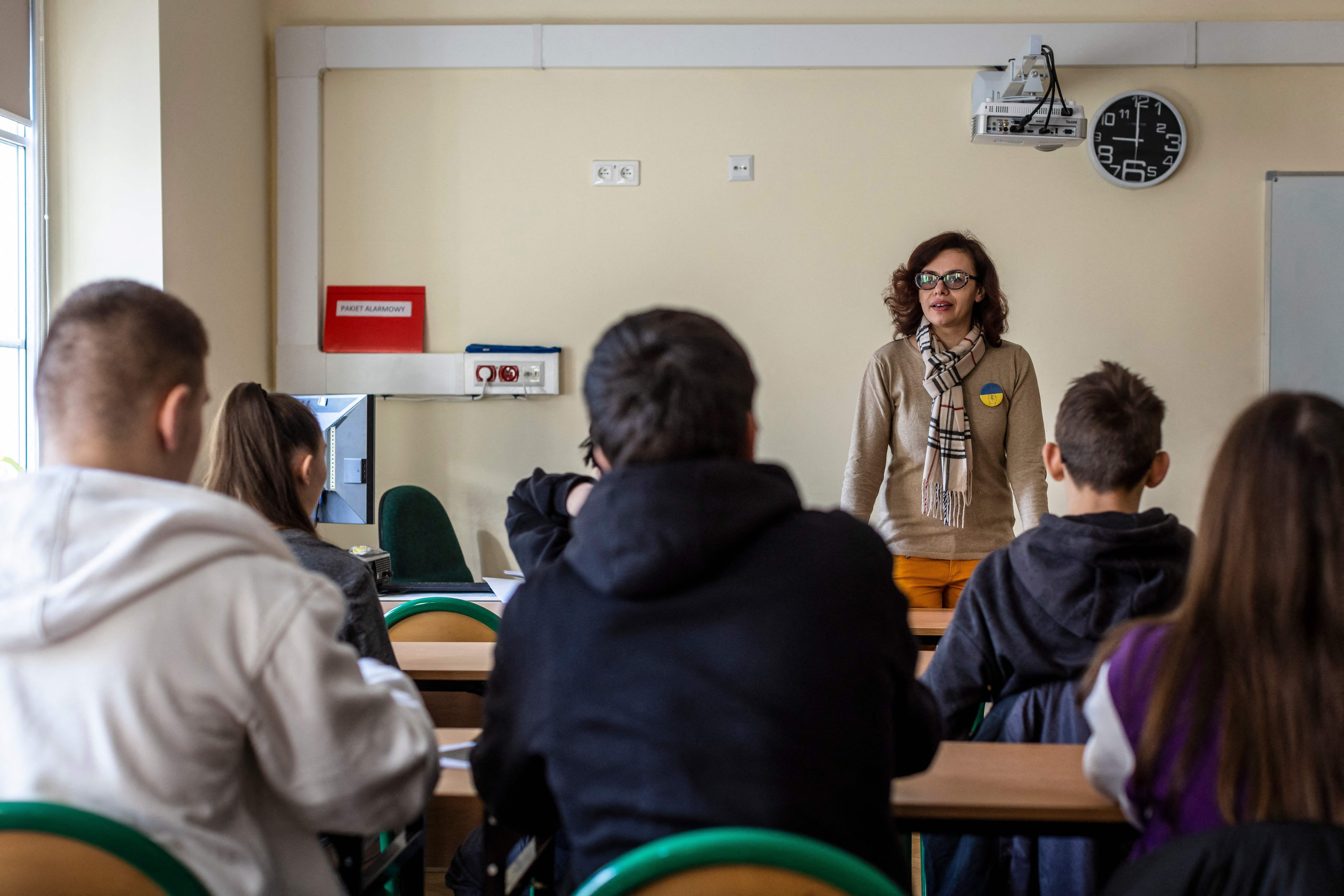 Teacher speaking; students seated; classroom with a clock, projector, and wall sign reading "PAKIET ALARMOWY."