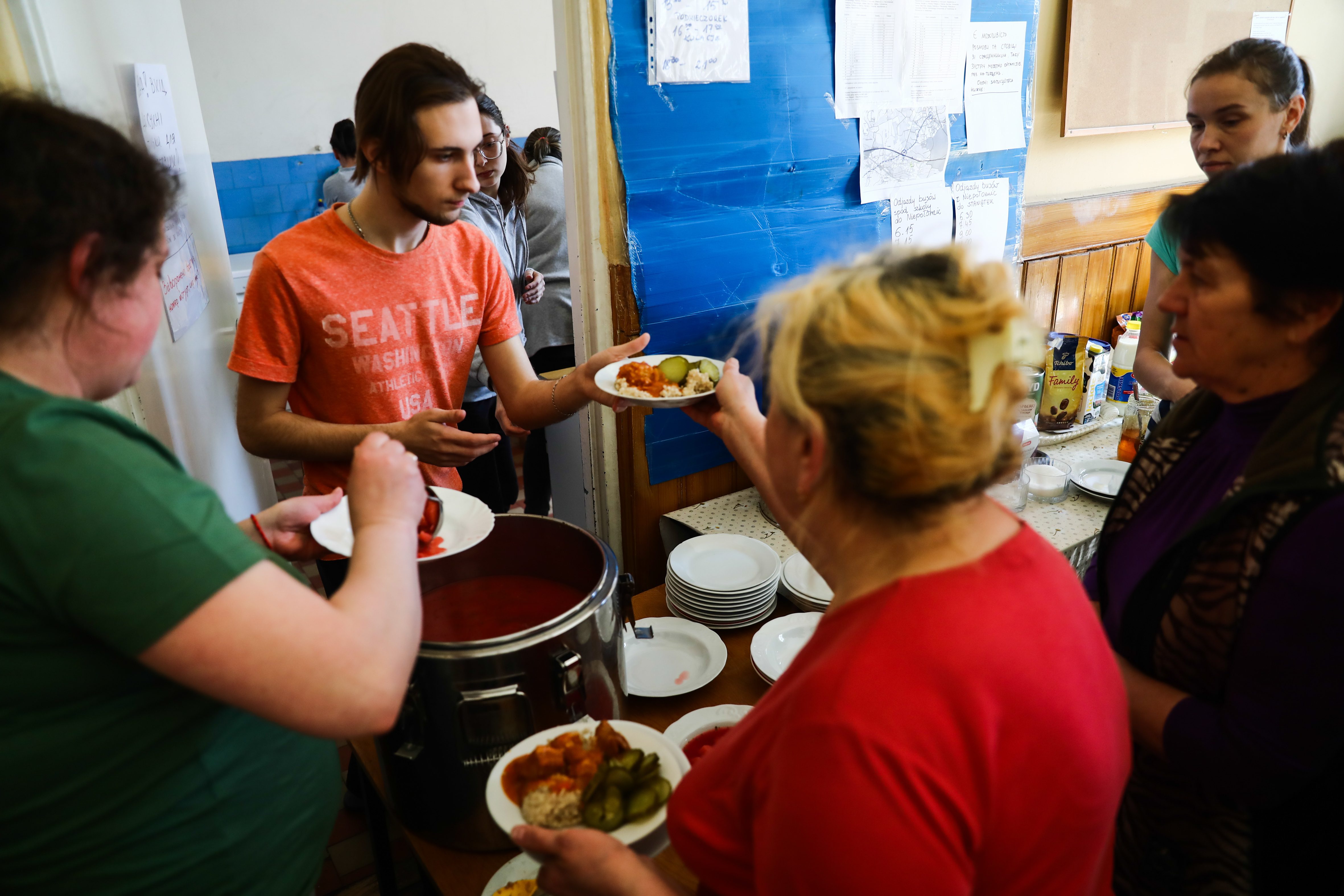 People serve food from a large pot, distributing plates in a community setting. Walls display papers and boxes are visible nearby.