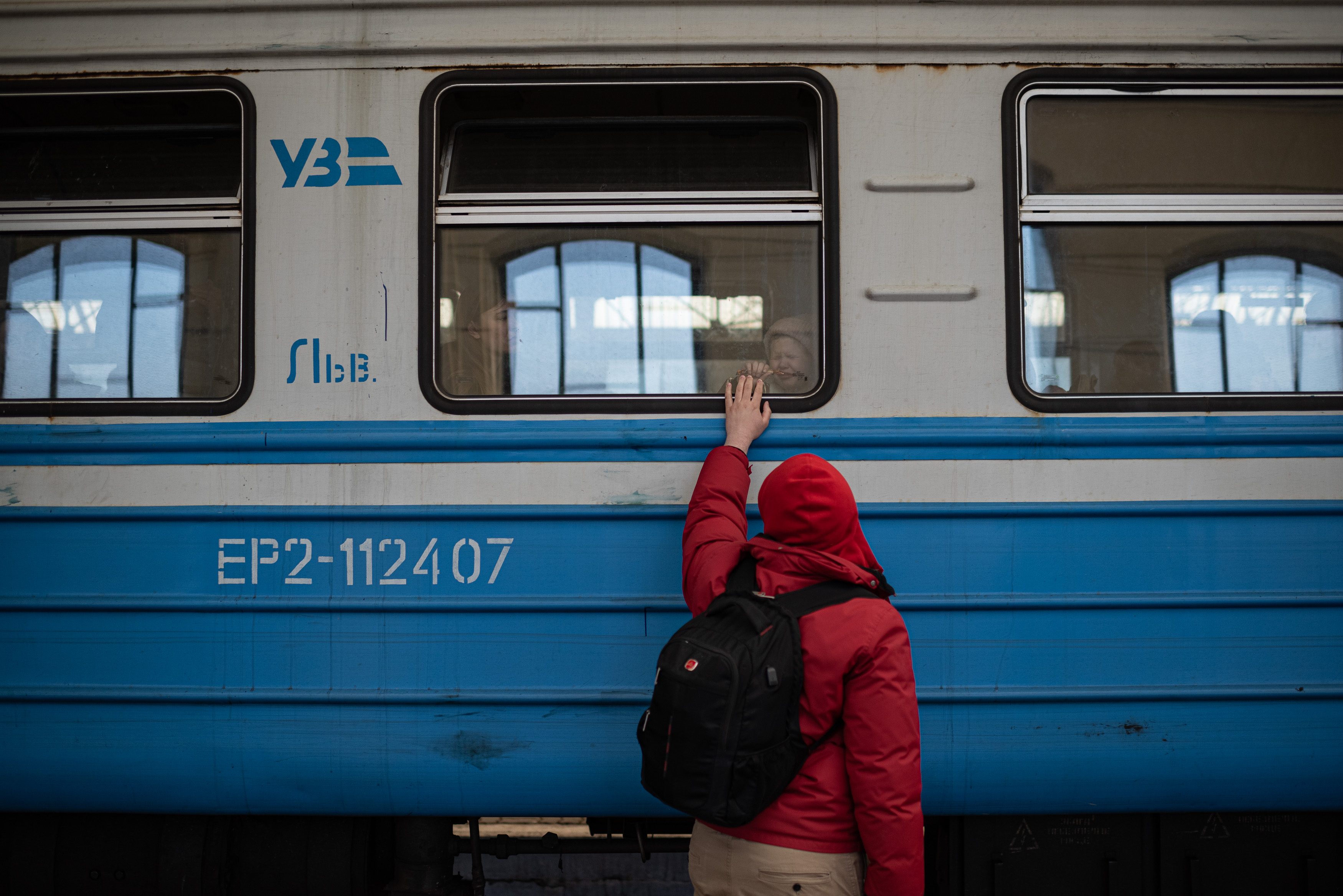 A person in a red jacket touches a train window, someone inside smiles back. The train has numbers "Ð•Ð 2-112407" and letters "Ð£Ð—".
