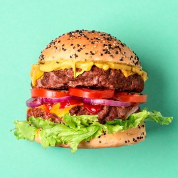 Burger displaying layers of beef, cheese, lettuce, tomato, and onions, sits against a solid teal background.