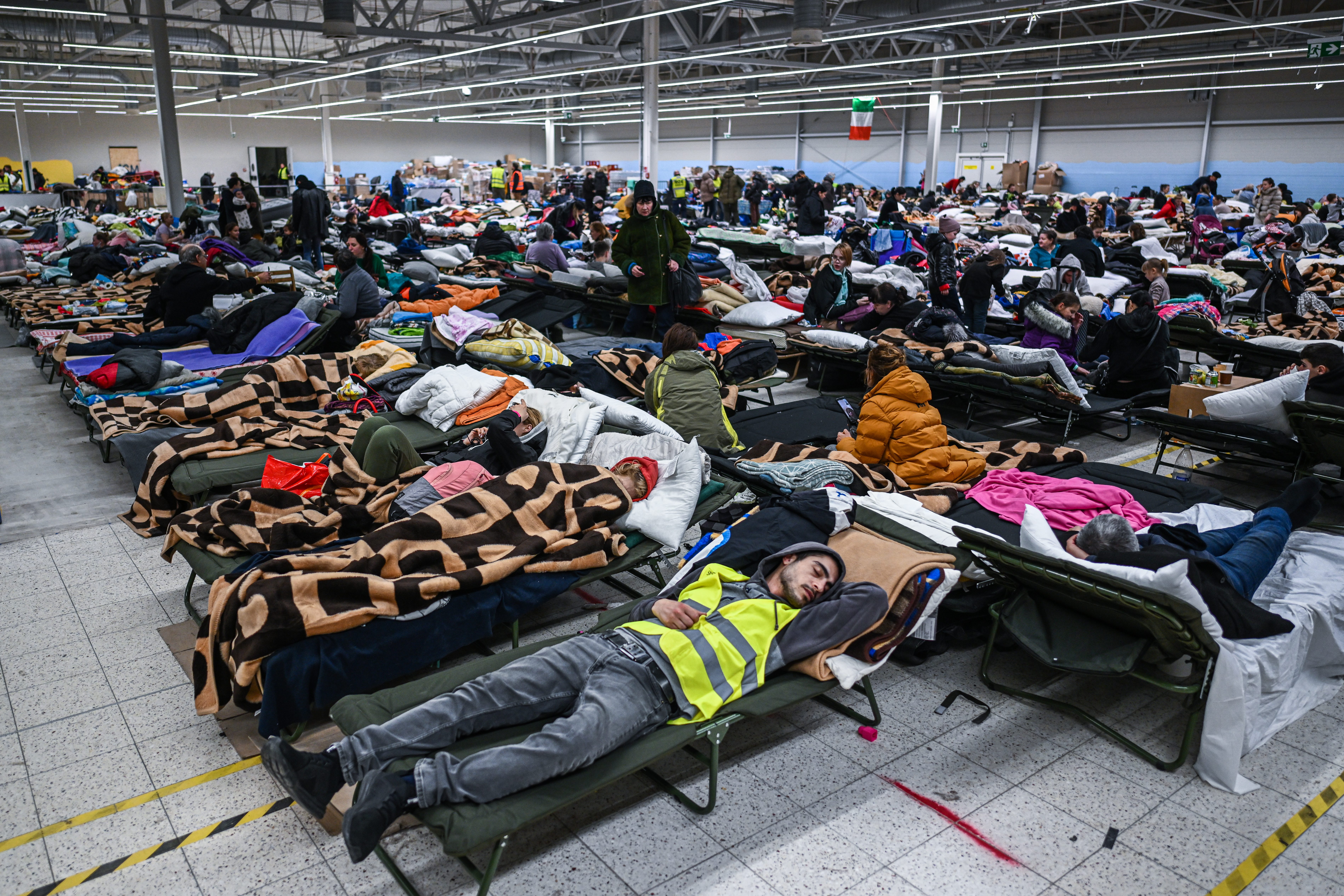People rest on cots, covered in blankets, in a large indoor shelter filled with numerous beds and belongings.