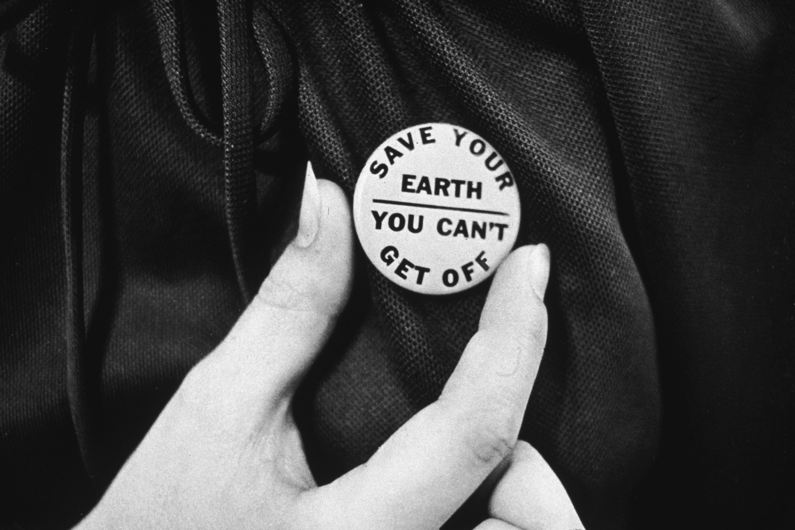 Hand holds button with text: "SAVE YOUR EARTH YOU CAN'T GET OFF," against a textured fabric background.