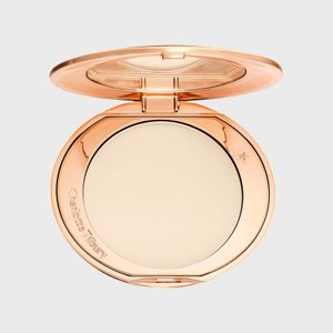 Compact powder sits in an open round, gold case, displaying a light beige shade. The lid reflects light against a plain gray backdrop.