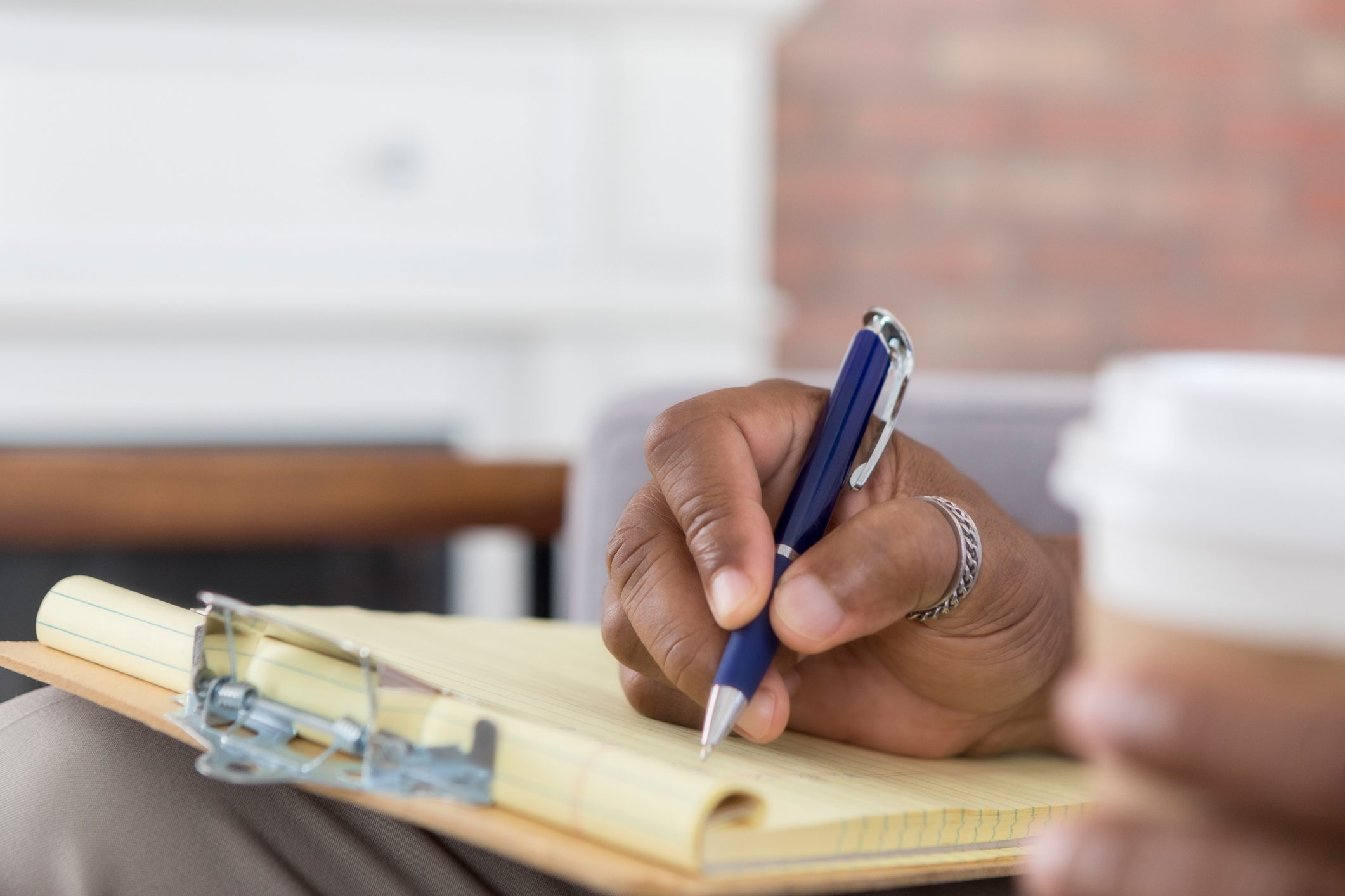 Hand holding a pen writes on a yellow notepad, with a coffee cup nearby, in a cozy indoor setting.