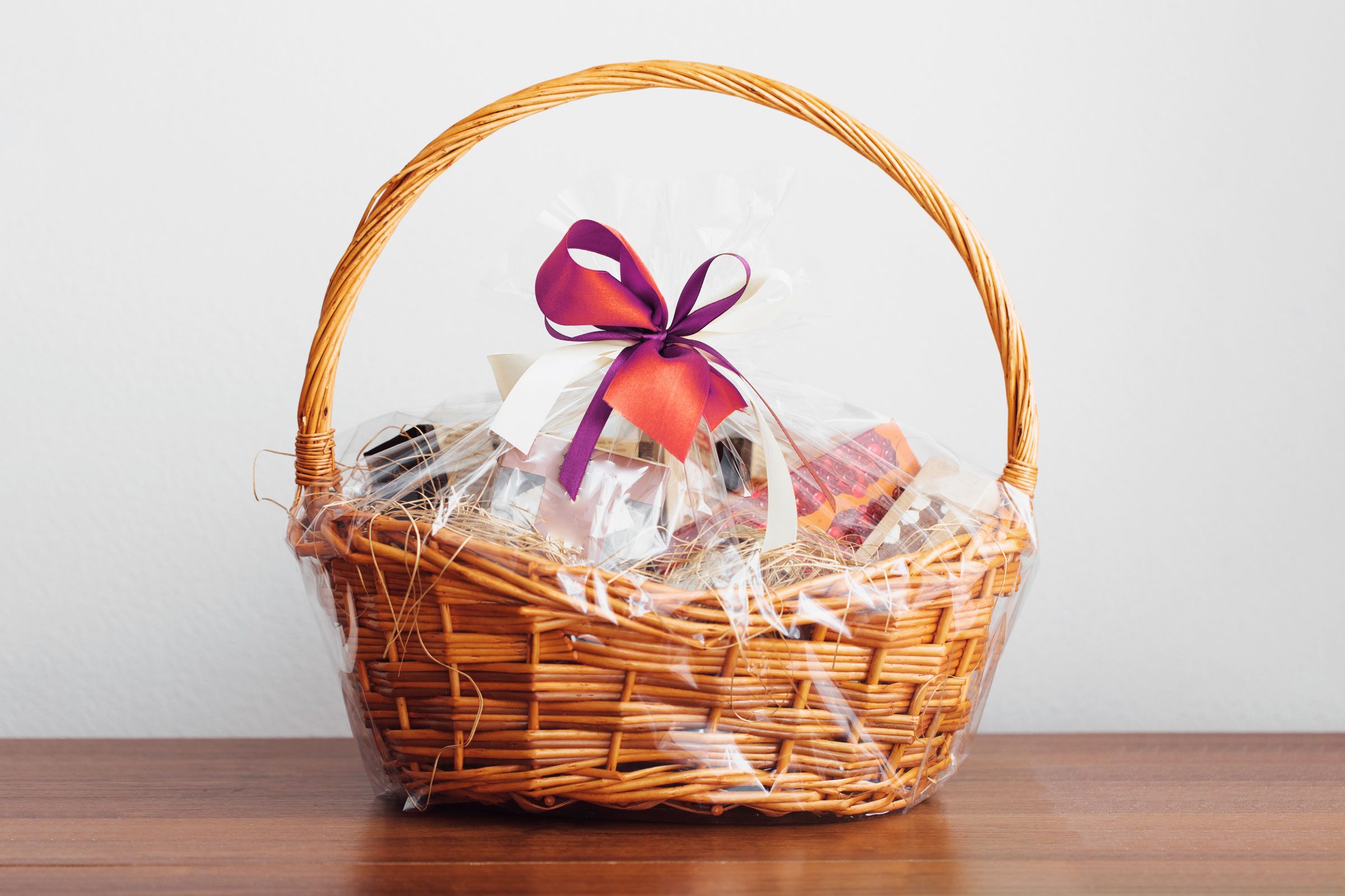 A wicker basket wrapped in transparent plastic contains assorted gifts, adorned with a purple and orange bow, sitting on a wooden table.