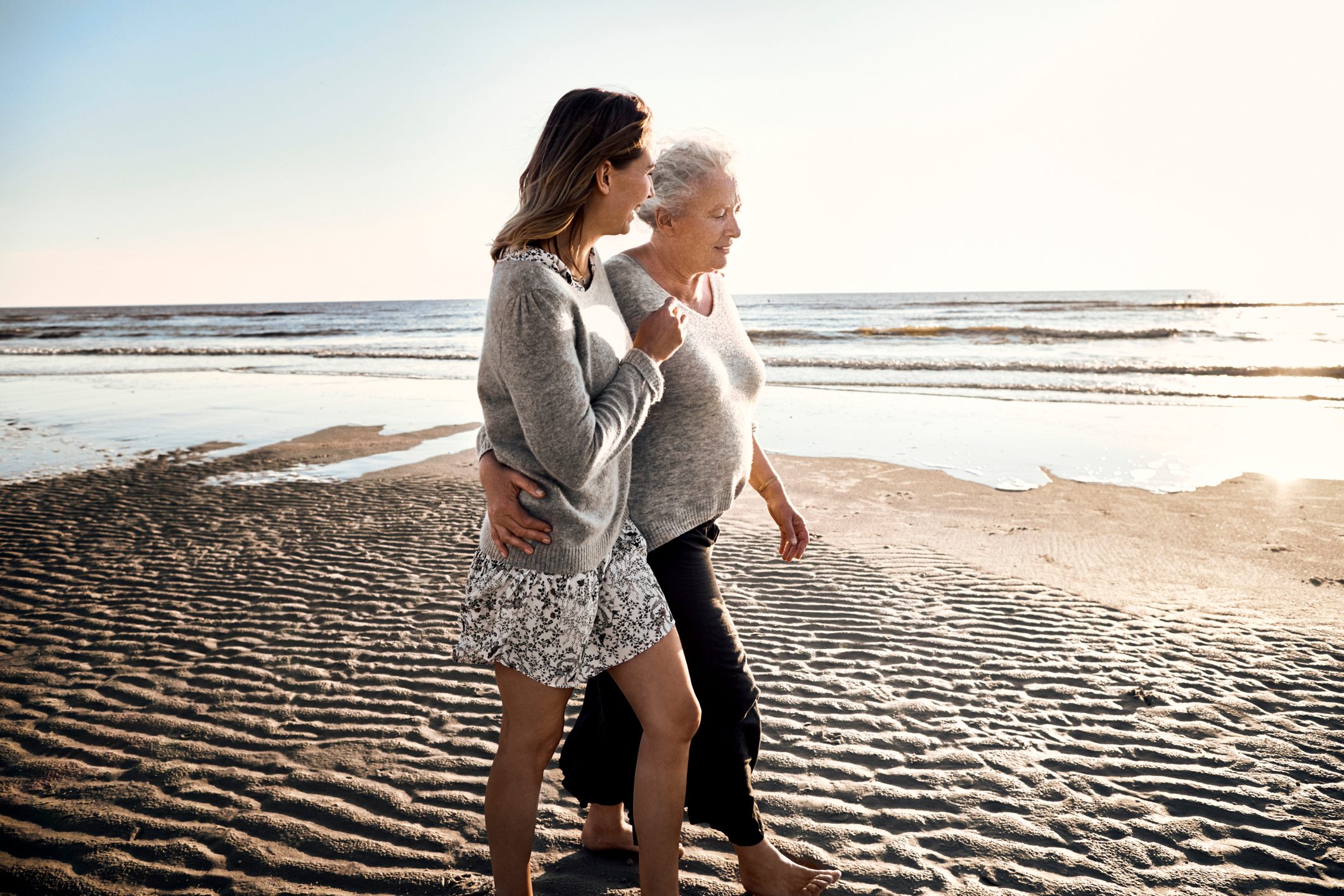 Two people walk together on a sandy beach, embracing warmly, with the ocean and a clear sky in the background.