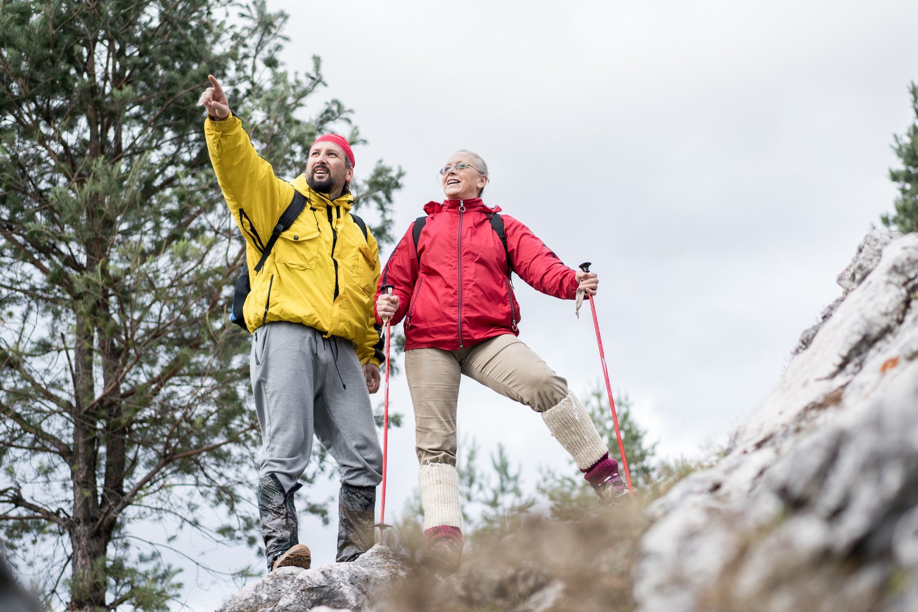 Two hikers, wearing colorful jackets, stand on rocky terrain pointing and smiling, surrounded by trees and an overcast sky.