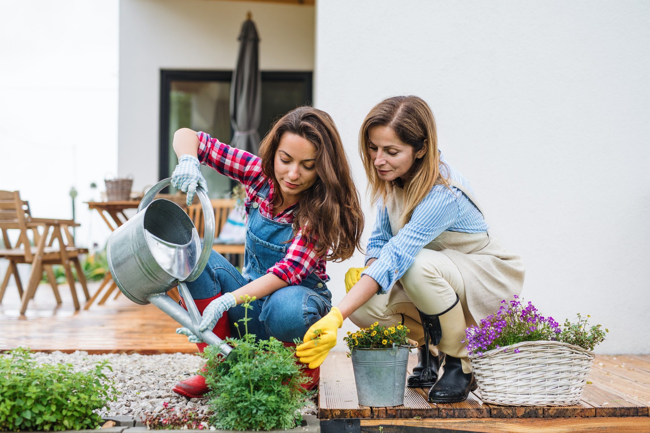 Two women are gardening; one waters plants, the other tends flowers. They're on a wooden deck beside a house with patio furniture.