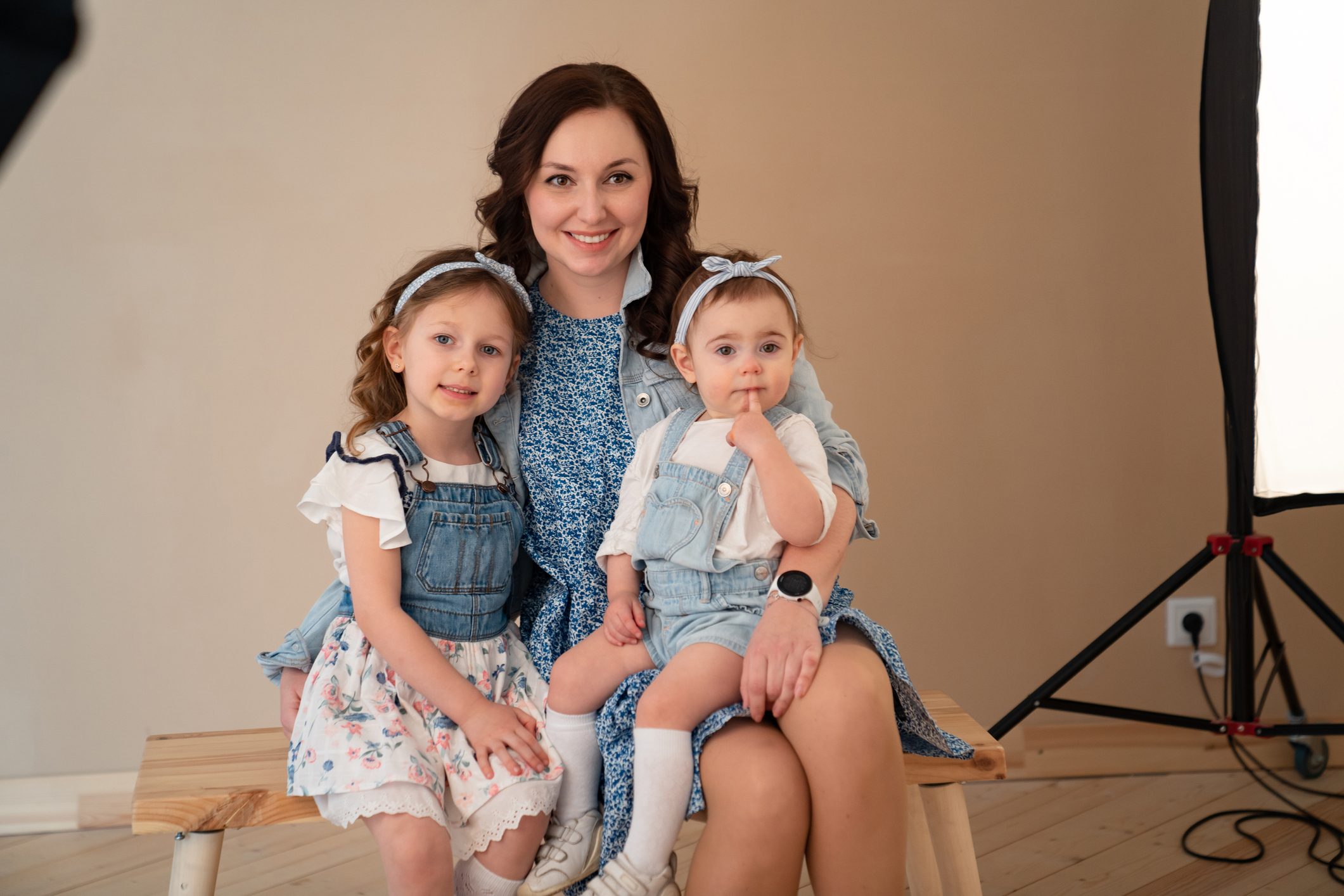 A woman and two children sit together, smiling, on a wooden bench in a well-lit photography studio with beige walls and wooden floor.