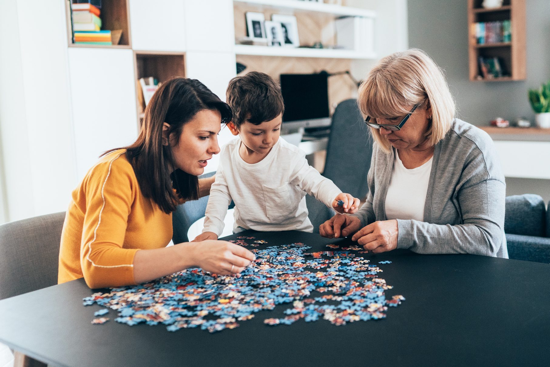 Three people assemble a jigsaw puzzle on a black table in a cozy living room with shelves and a computer in the background.