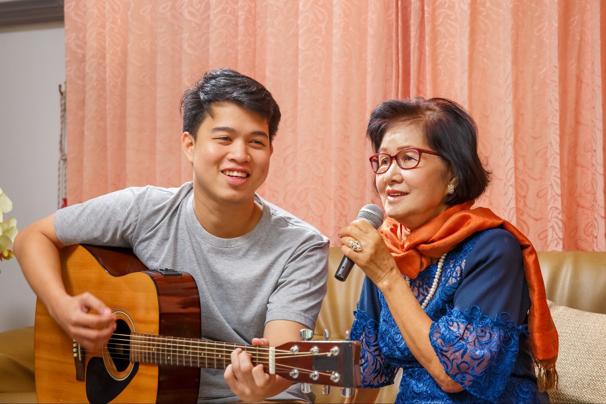 Two people perform music on a couch; one plays a guitar, the other sings into a microphone, surrounded by patterned curtains.