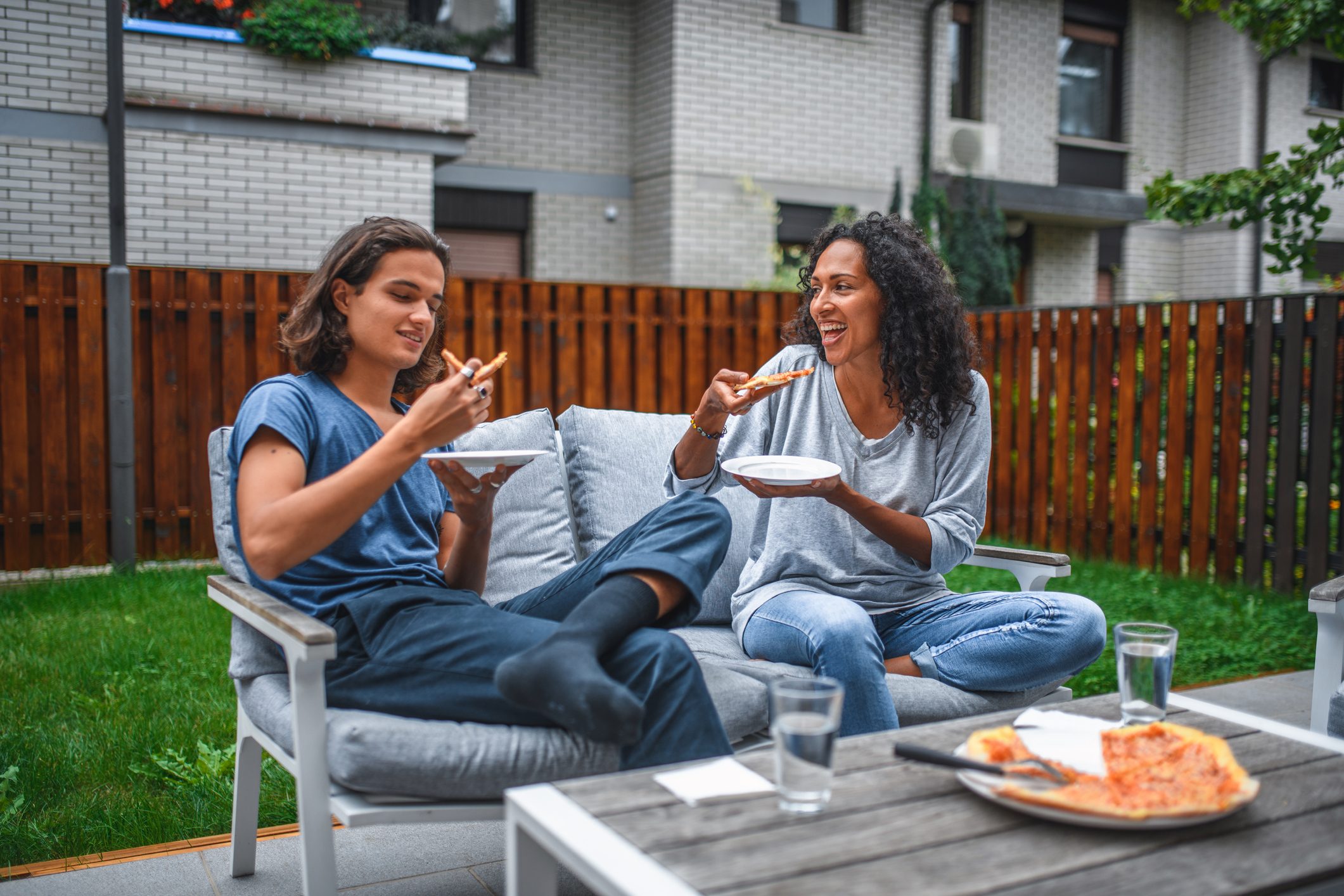 Two people eat pizza, seated on a patio couch, surrounded by a wooden fence and green lawn.