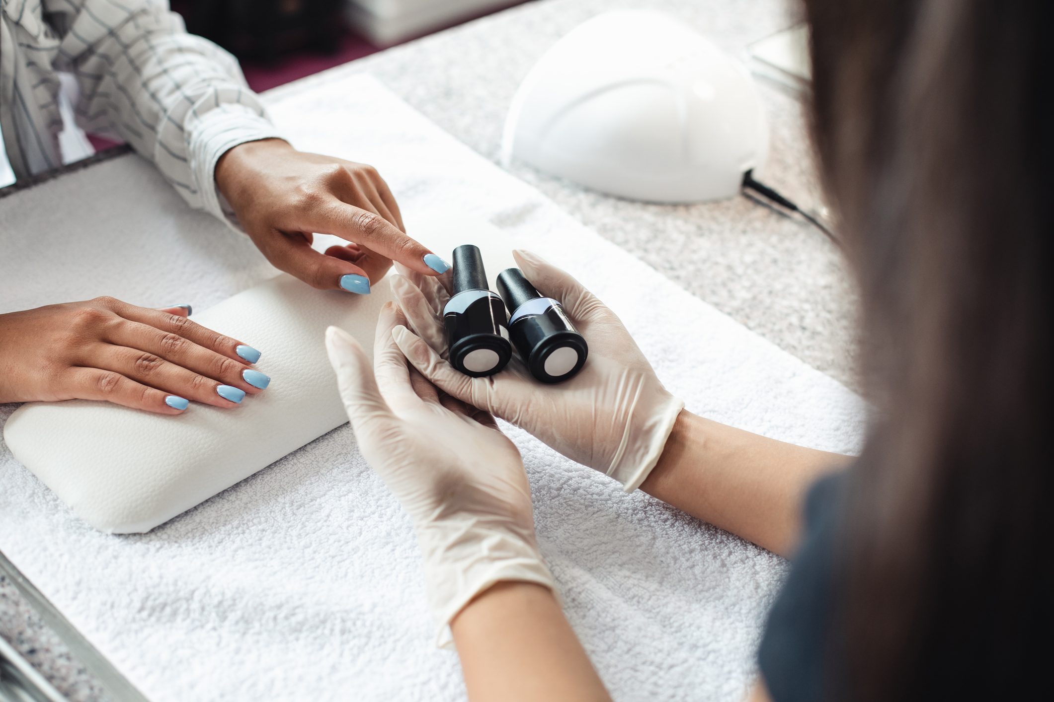 Hands choosing nail polish bottles; wearing gloves in a manicure salon on a white towel-covered surface.