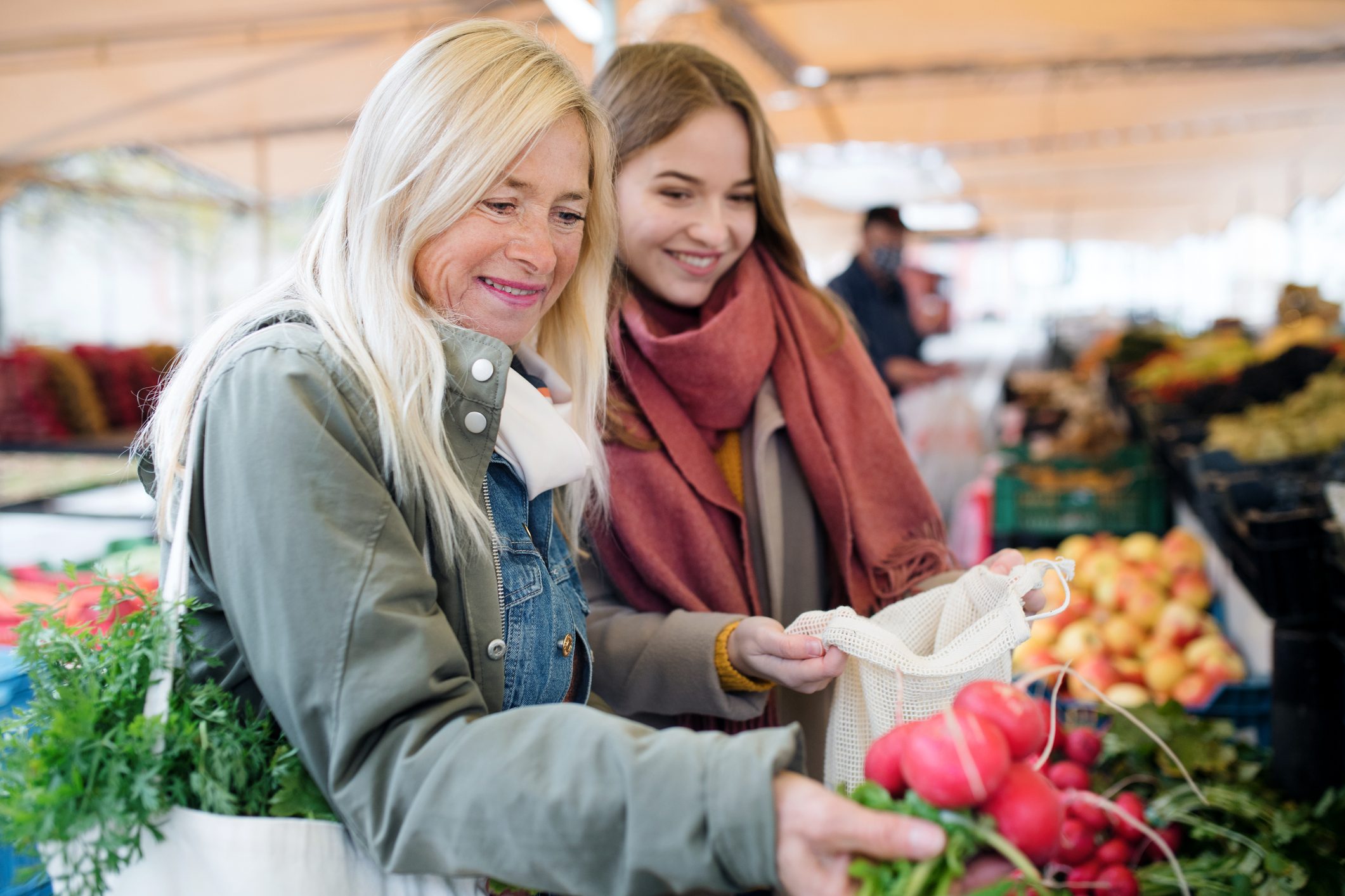 Two smiling individuals select vegetables at a bustling outdoor market, surrounded by fresh produce in crates and colorful stalls.