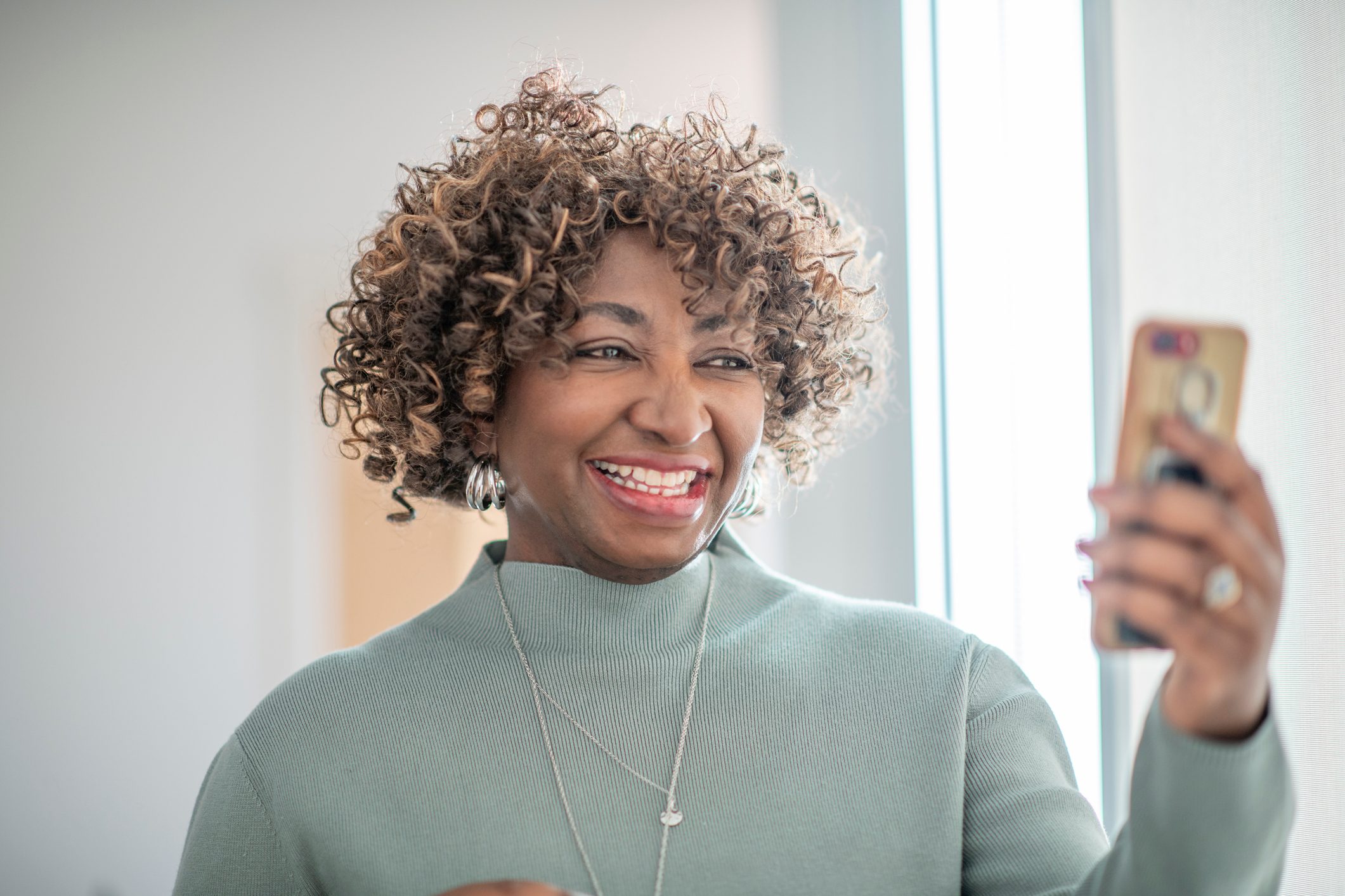 Woman smiling, holding a smartphone, indoors with bright natural light from a nearby window.
