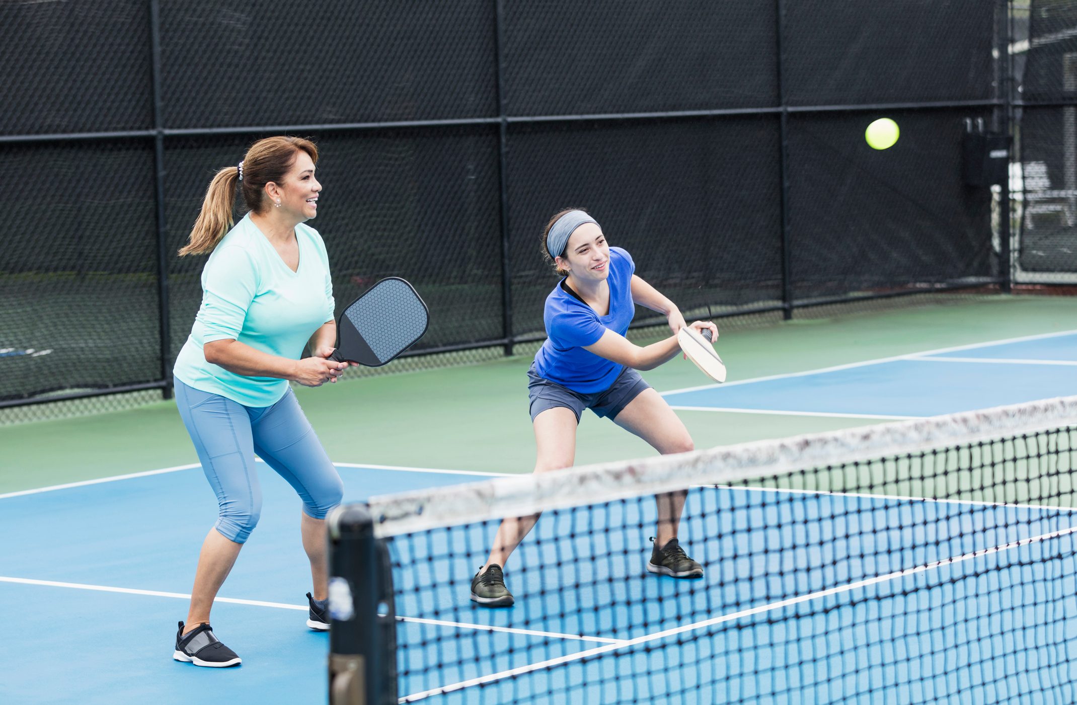 Two people play pickleball on a court, one hitting the ball, the other preparing, with a net separating them from the opponent's side.