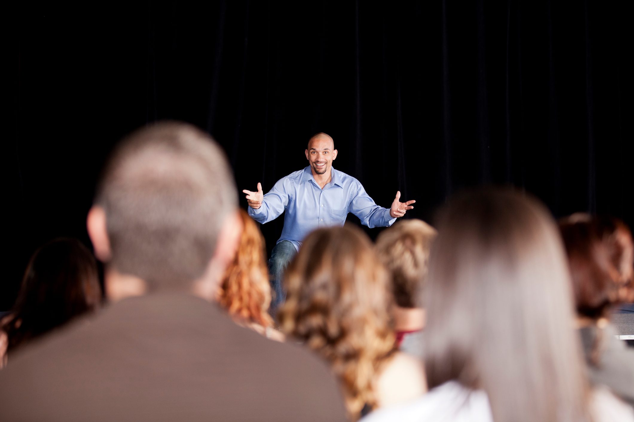 Speaker gestures enthusiastically while addressing an audience, seated in a dark theater setting.