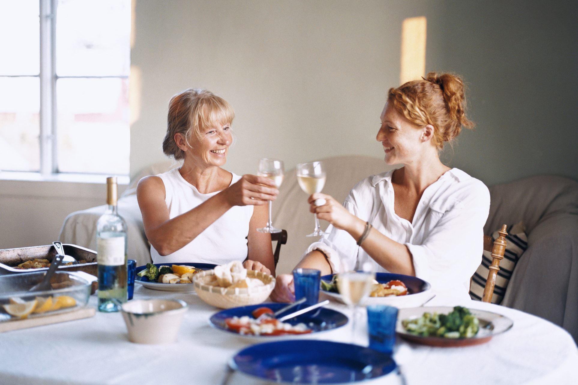 Two people clink wine glasses, smiling, at a dining table filled with food in a softly lit room.