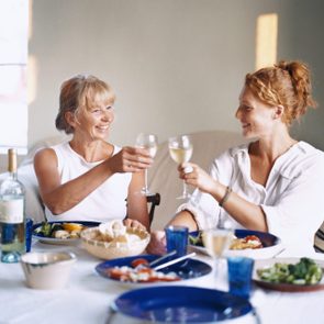 Two people clink wine glasses, smiling, at a dining table filled with food in a softly lit room.