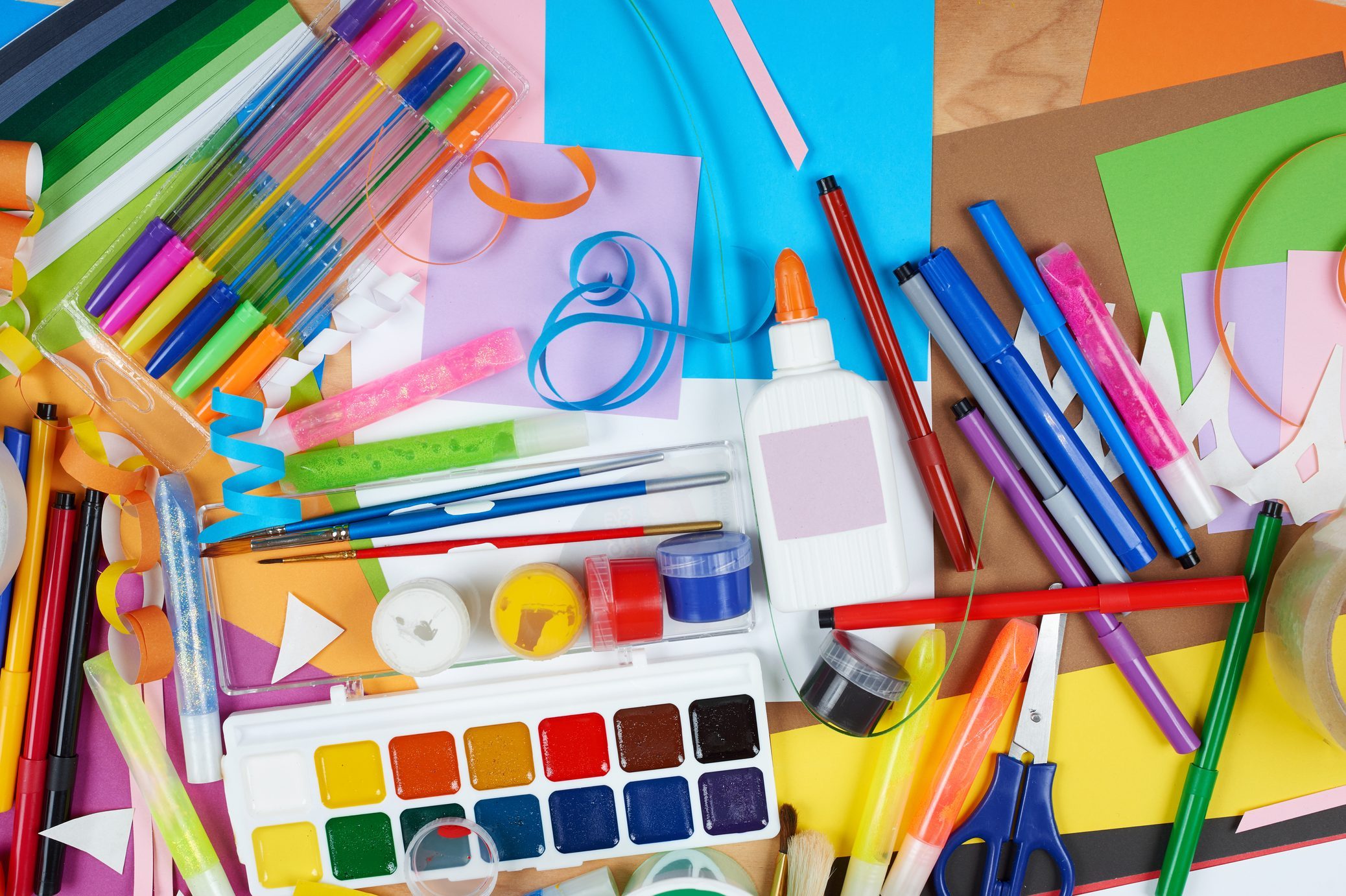 Art supplies scattered on colorful paper include markers, pens, scissors, paint palette, glue, jars, and ribbons, creating a vibrant crafting scene.