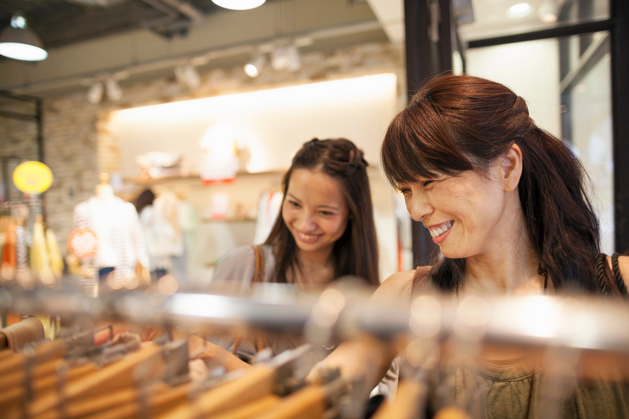 Two women smile while browsing clothes on hangers in a brightly lit store with blurred shelves in the background.