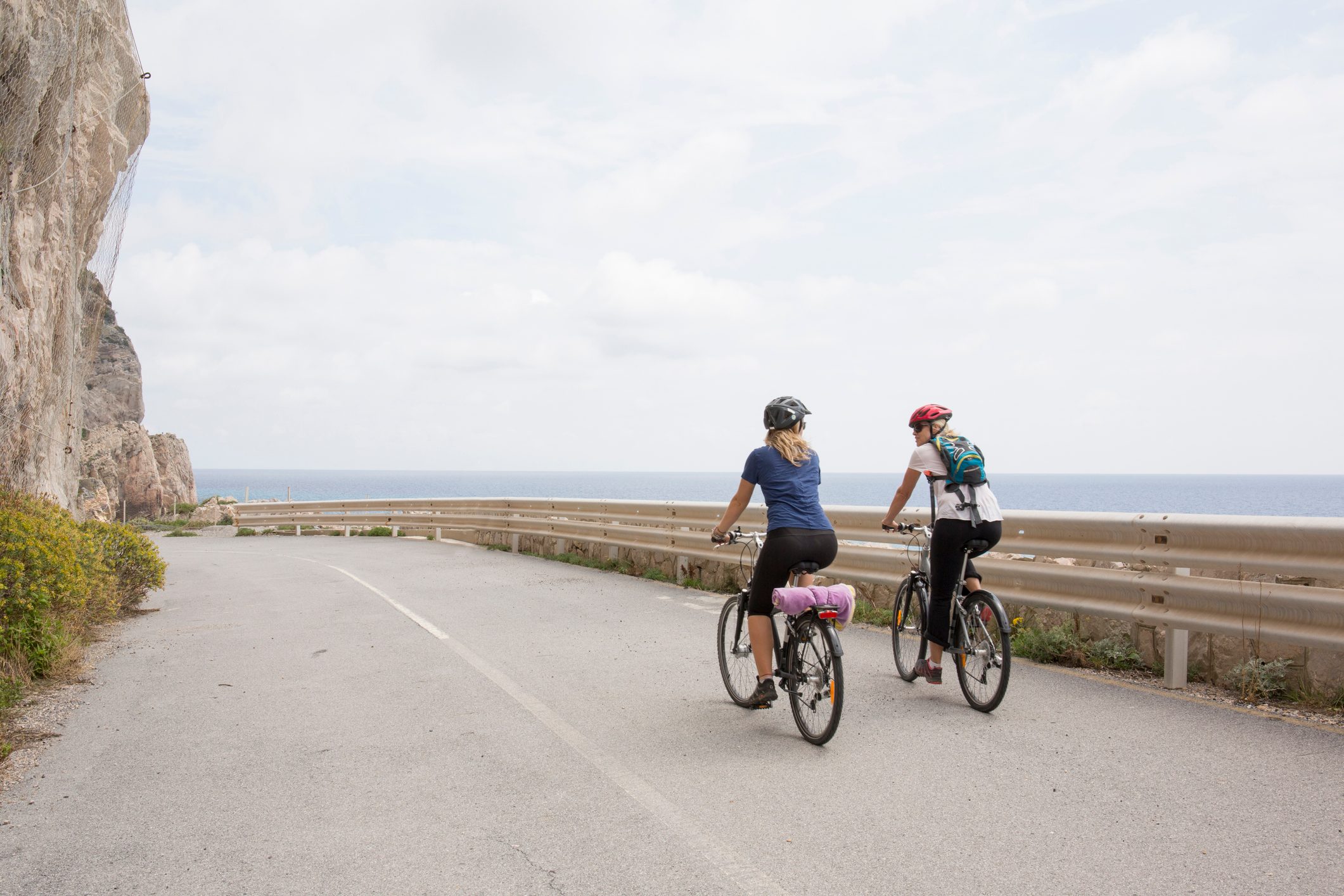 Two cyclists ride along a coastal road, with a rocky cliff to the left and the ocean in the background.