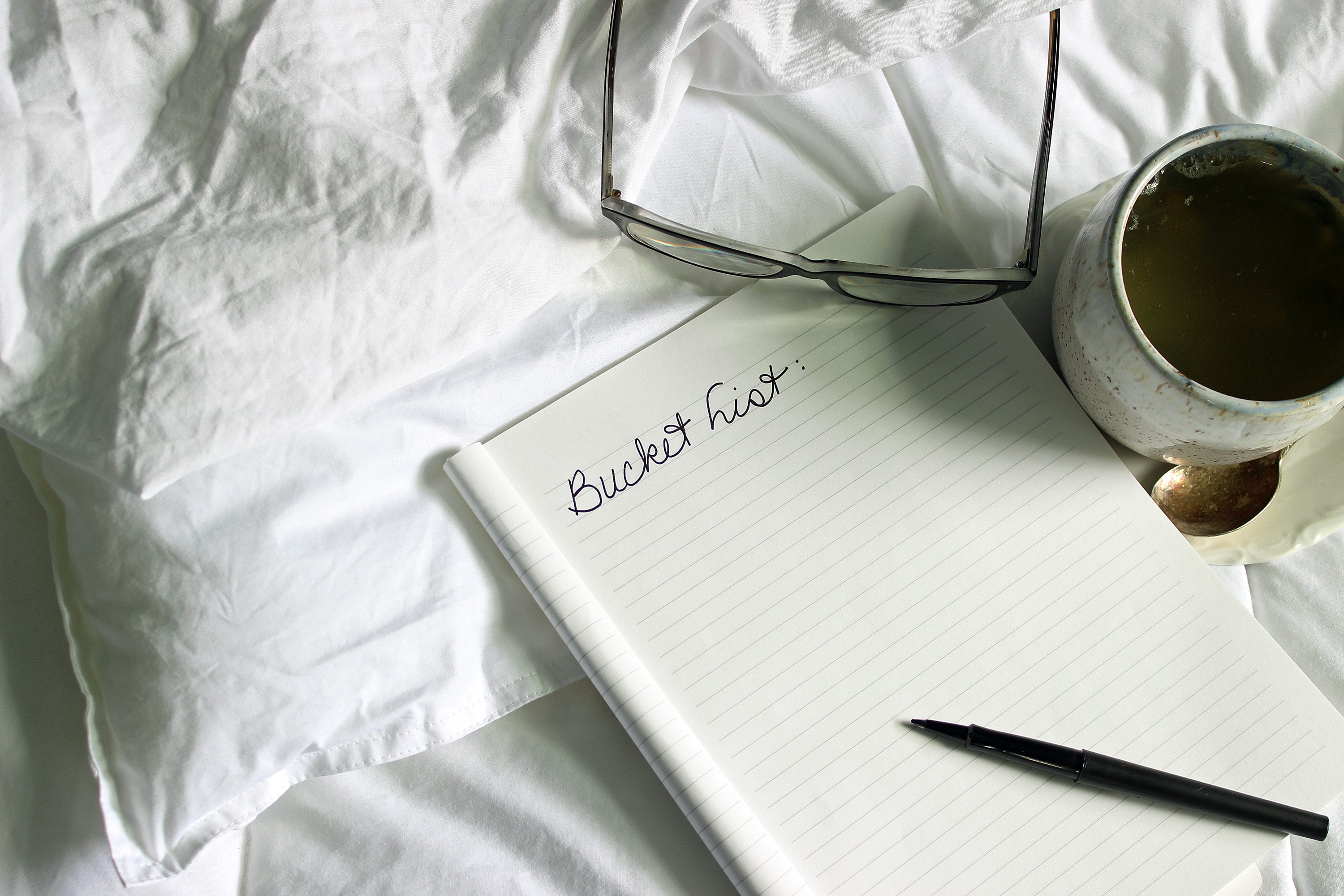 A notebook lies open on rumpled sheets, titled "Bucket List," with a nearby pen, glasses, and a cup on a saucer.