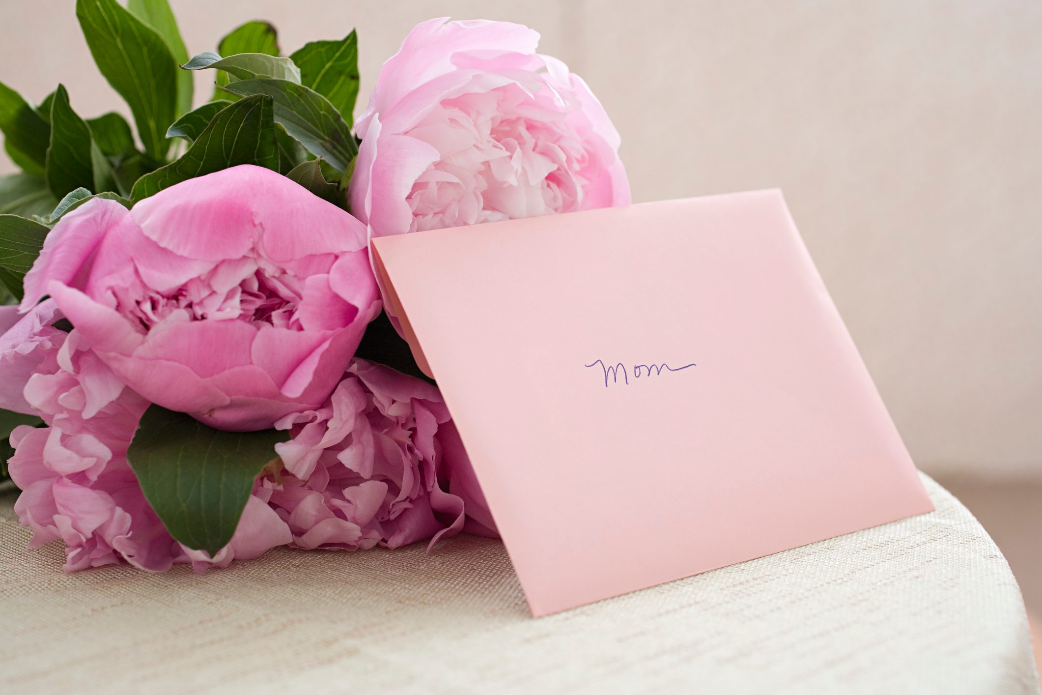 Envelope labeled "Mom" rests against pink peonies, placed on a light-textured surface.