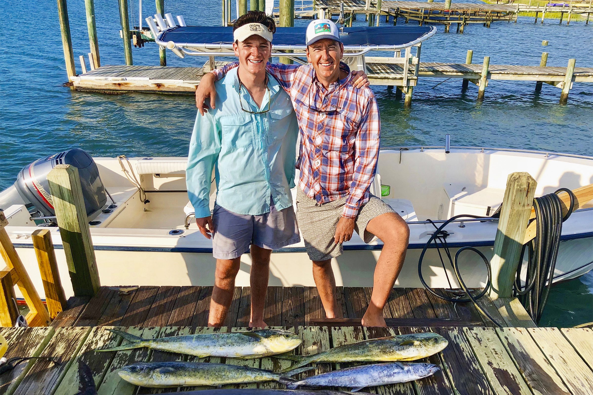 Two people smile on a dock beside a boat, displaying freshly caught fish, with a marina in the background under a sunny sky.