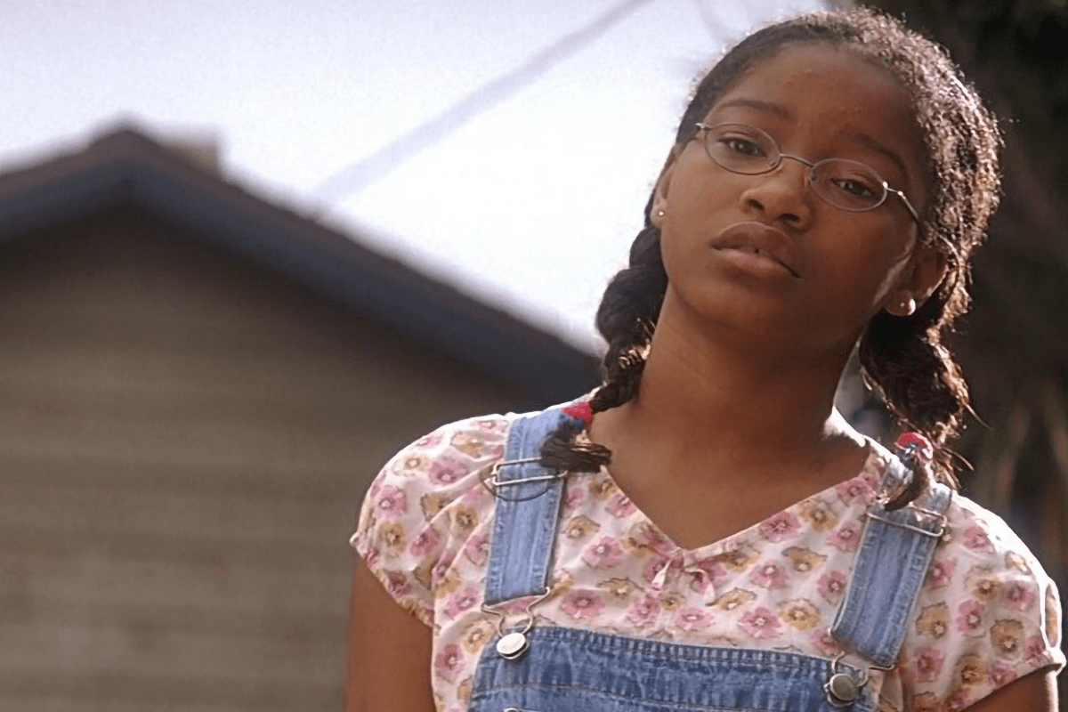 A young girl gazes intently, wearing glasses and floral shirt with denim overalls, set against an outdoor background with a blurred house.