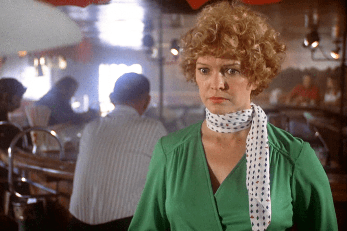 A person stands, looking serious, wearing a green outfit with a white polka-dot scarf in a bustling diner setting.