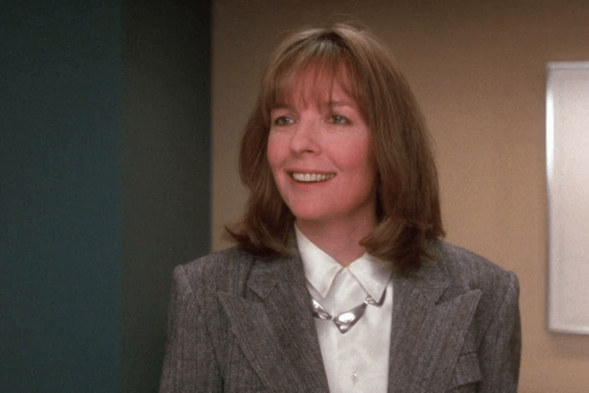 A woman smiles while standing, wearing a gray blazer and white shirt, in an indoor setting with teal and beige walls.