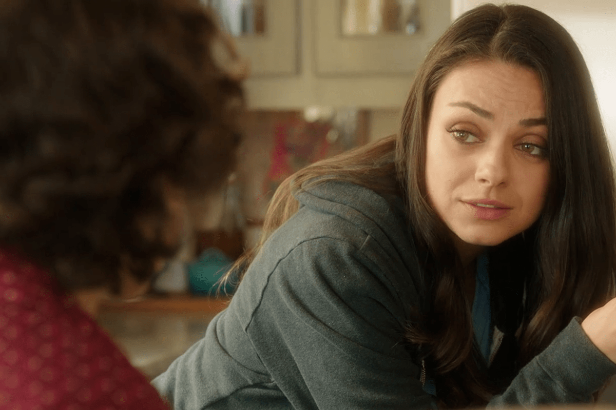 A woman leans on a surface, conversing with a person wearing a red patterned shirt in a well-lit, cozy room.