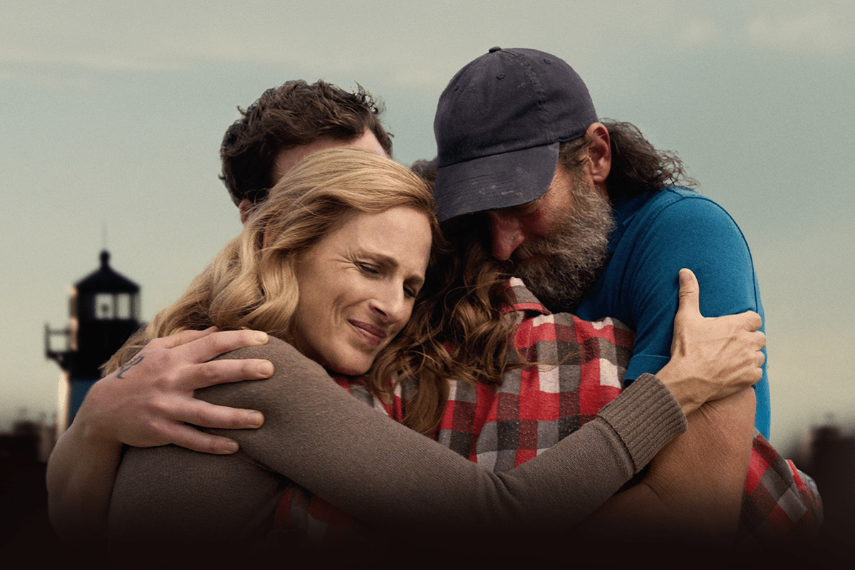 Four people embrace tightly outdoors, against a blurred background featuring a lighthouse.
