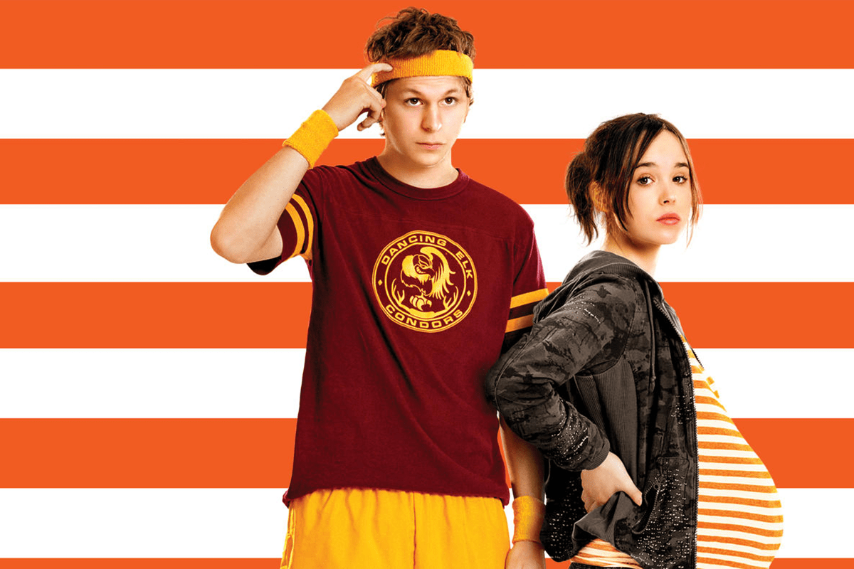 Two people stand side by side; one wears sports attire, while the other is pregnant, with an orange striped background displaying "Dancing Elk Condors."
