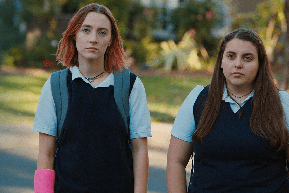 Two teenagers stand on a grassy street, one with a pink cast and backpack, wearing school uniforms.