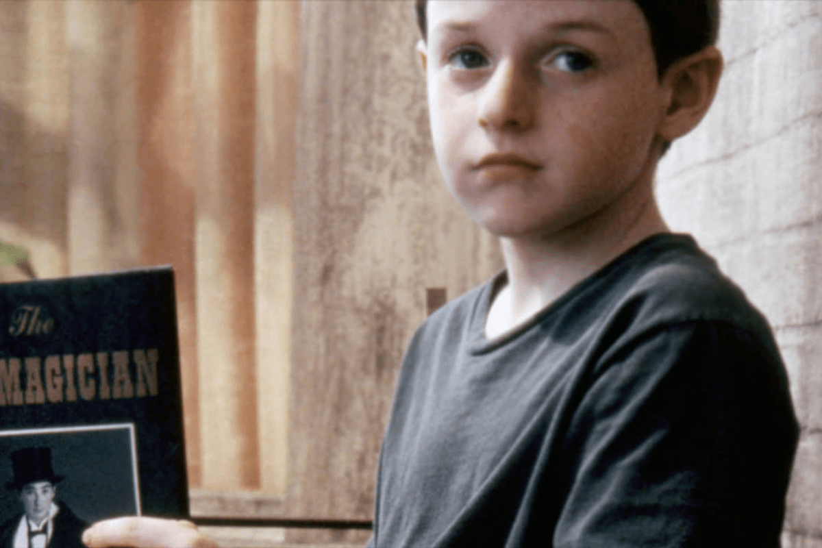 Child holding a book titled "The Magician," seated indoors against a softly lit, wooden wall.