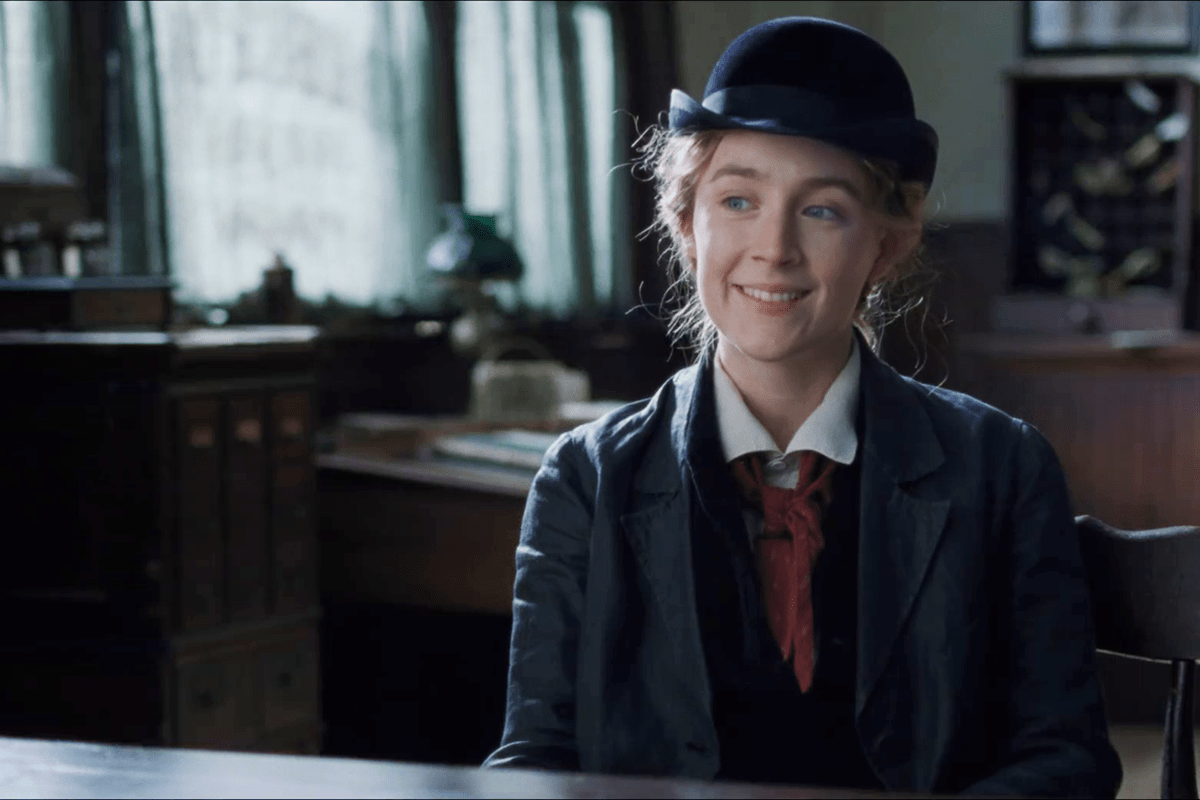 Smiling person sits at a desk, wearing a vintage suit with red tie and black hat, in a softly lit, traditional office setting.
