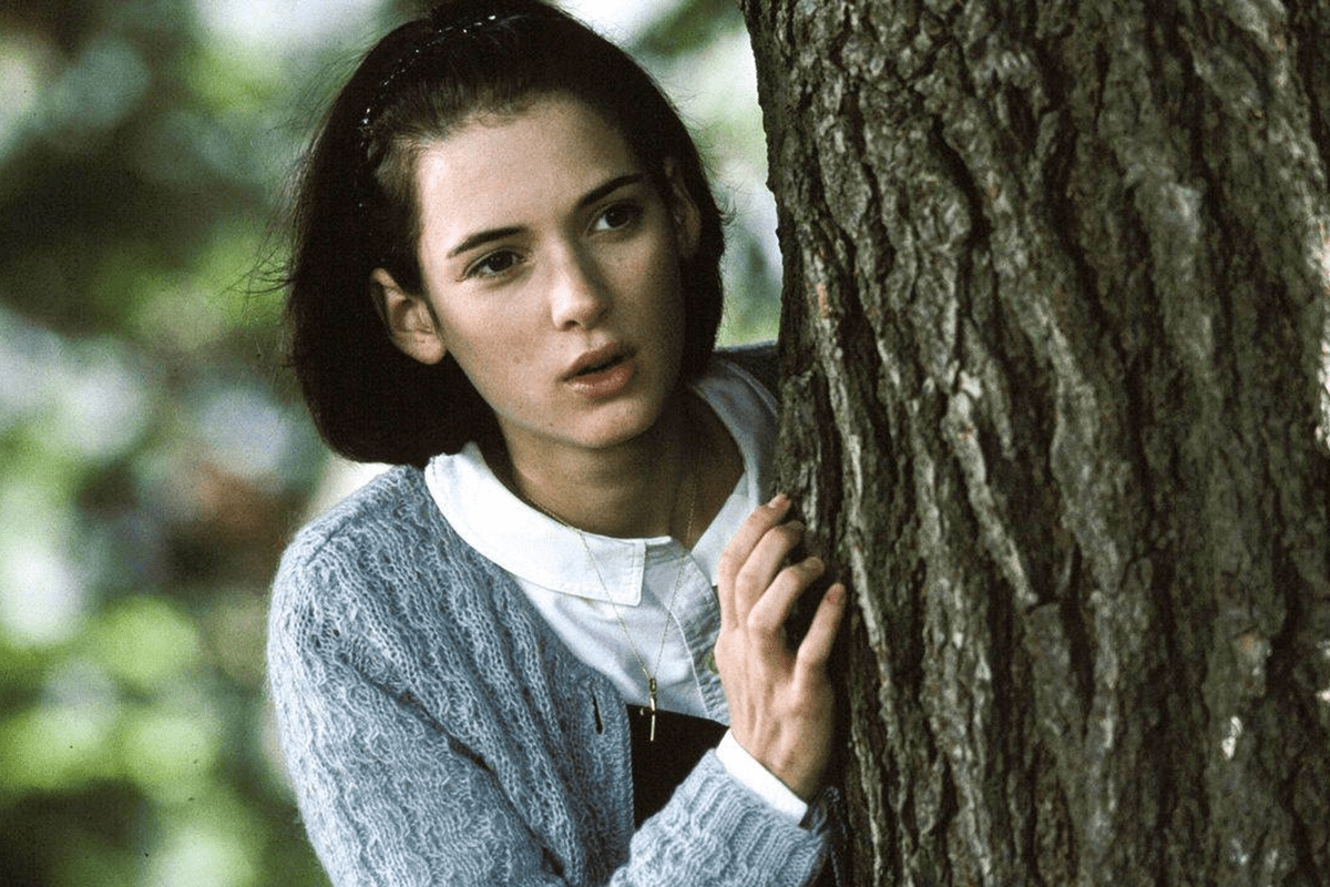 Person peering from behind a tree, wearing a blue sweater and white collared shirt, in a blurred, leafy outdoor environment.